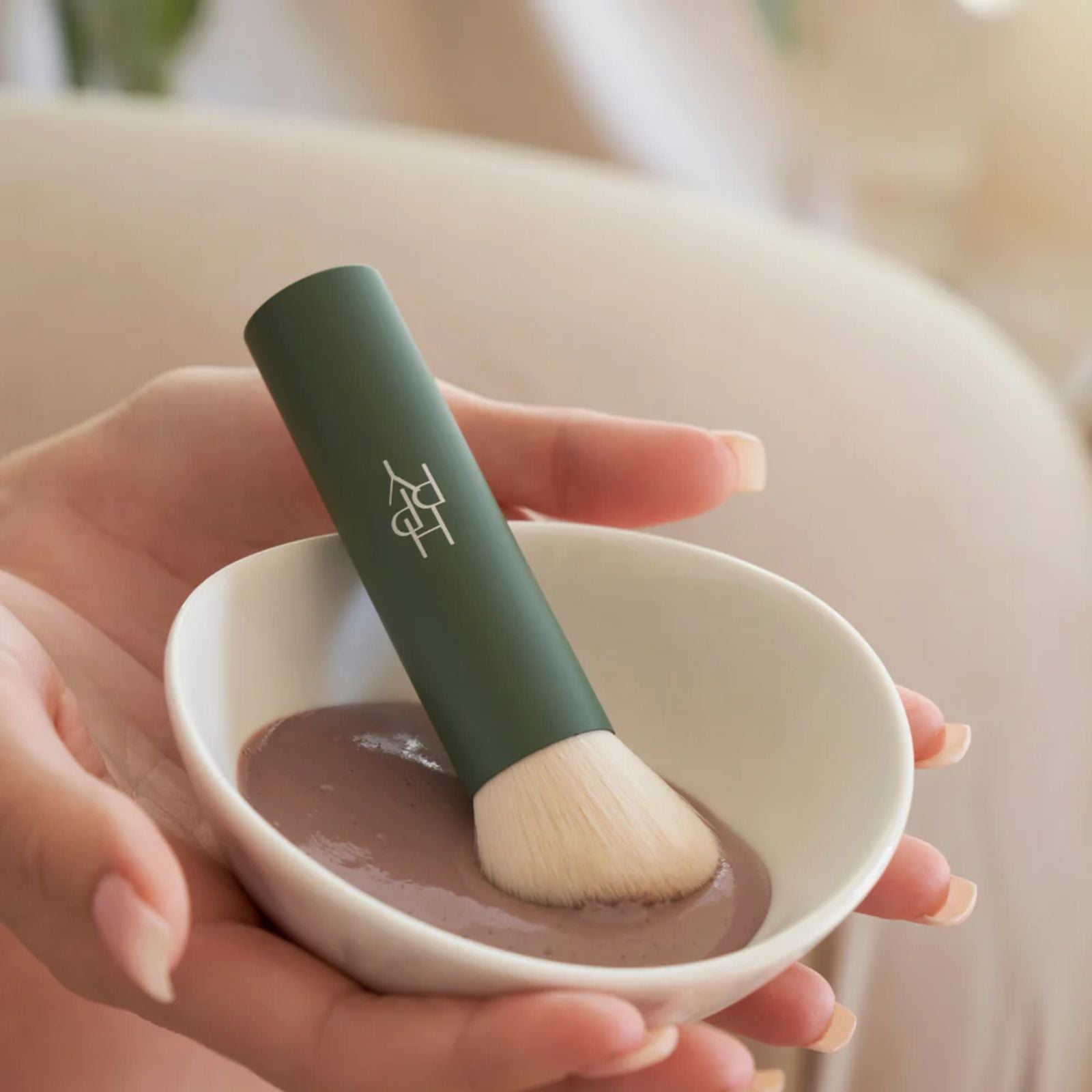 Hand holding a green makeup brush over a small white bowl with brown powder.
