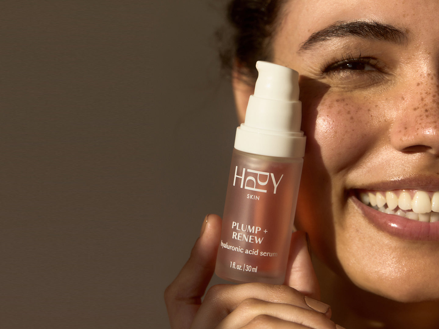 Woman holding a skincare serum bottle labeled 'HPPY' close to her face against a neutral background