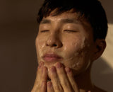 Person applying a facial mask with their hands against a neutral background