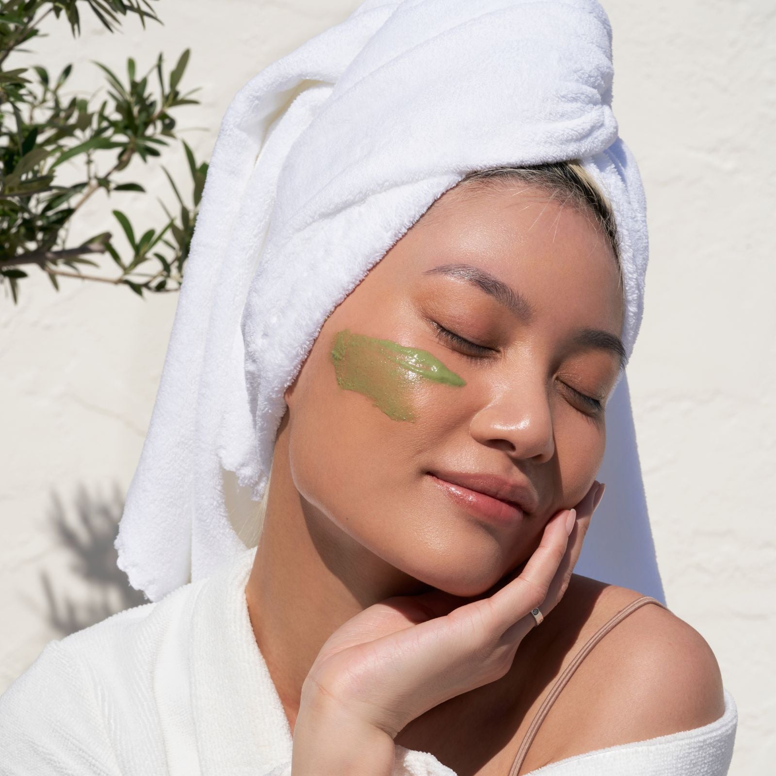Woman with a towel on her head applying green cream to her face outdoors.