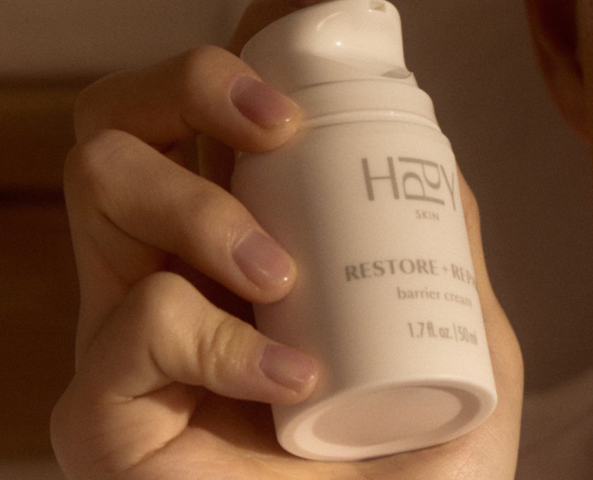 Hand holding a white skincare bottle labeled 'HPPY' with text on a blurred background