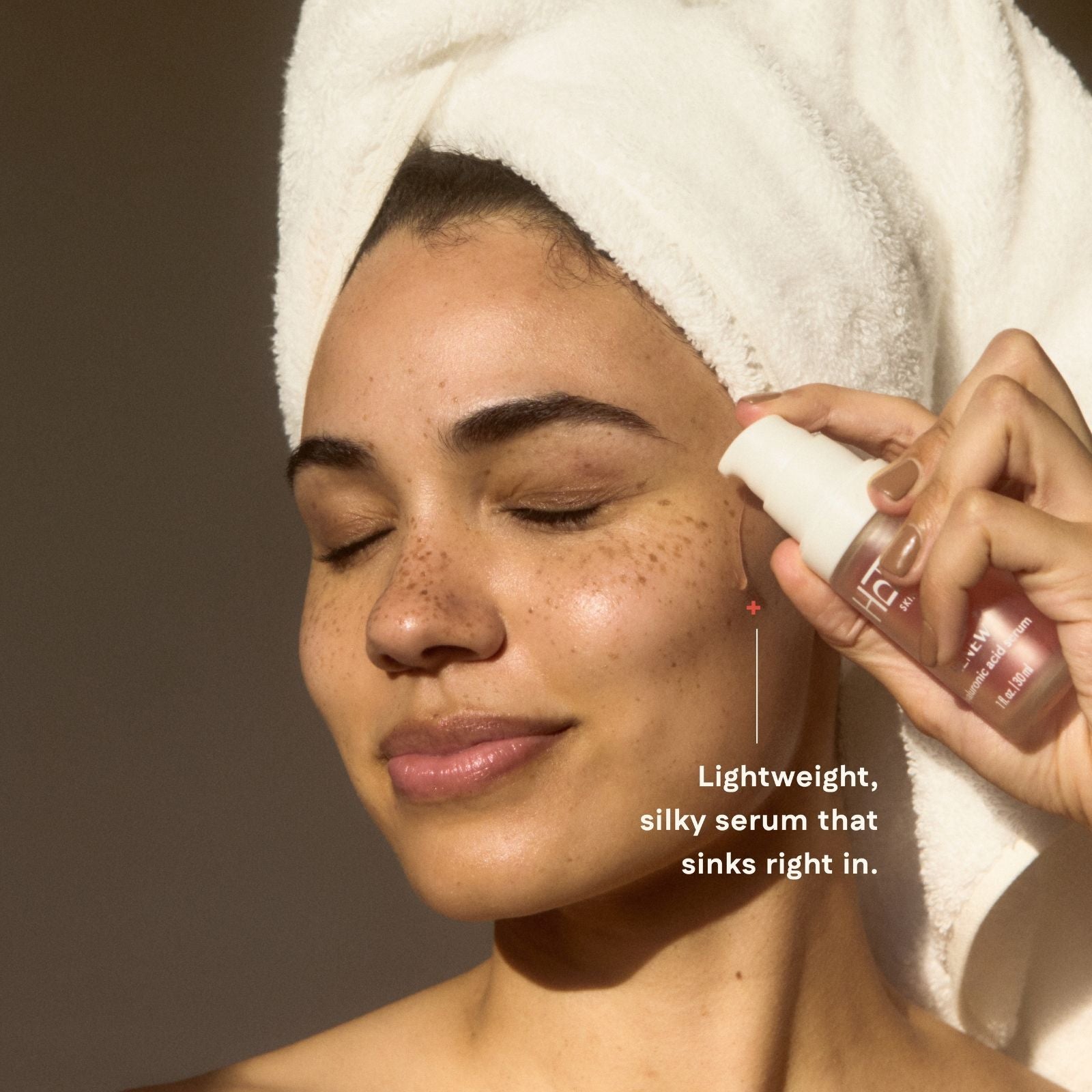 Woman applying a serum to her face with a towel on her head, against a neutral background.