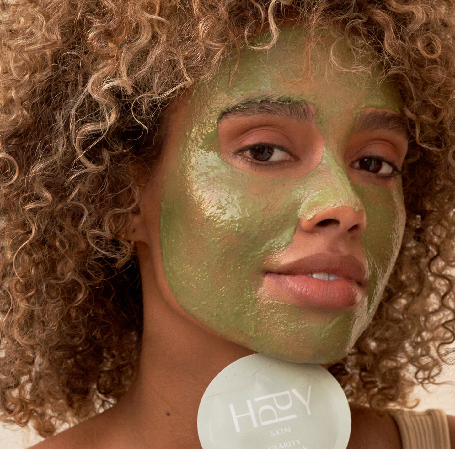 Woman with a green facial mask applying it to her face, holding a 'HARV' product.